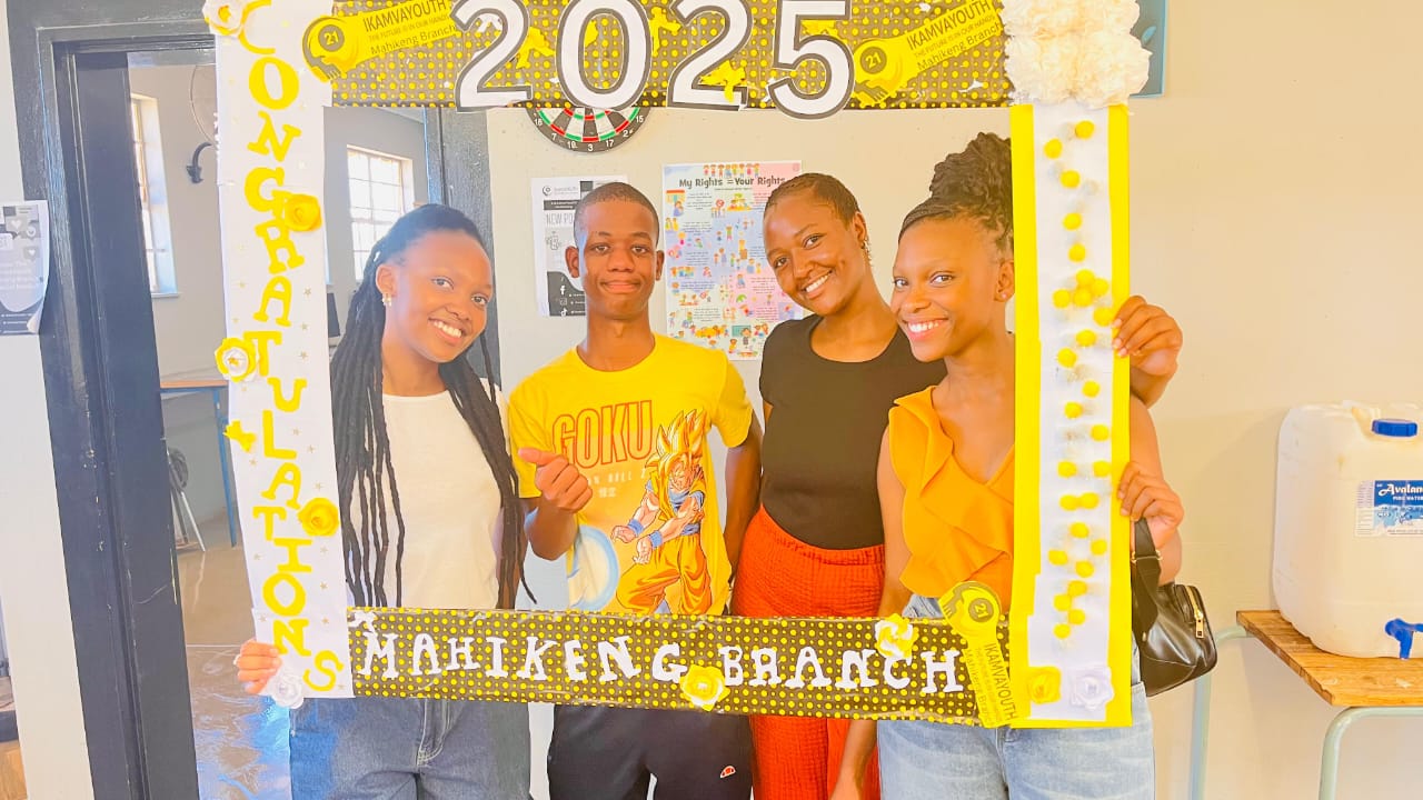 Four young people (one male, three female) in celebratory attire smiling and posing behind a large, decorated white and yellow "Congratulations" photo frame that reads "Mahikeng Branch" at the bottom.